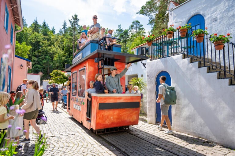 Trikken kjører rundt full av gjester i Kardemomme by i solskinnet.